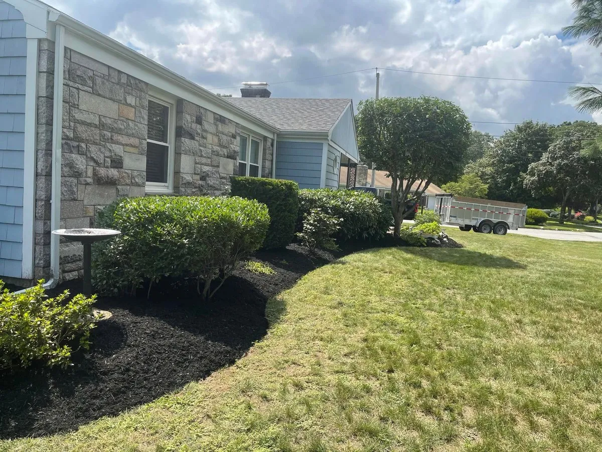 Fresh black mulch bed and trimmed shrubs along a stone and blue sided house, with a dump trailer in the driveway