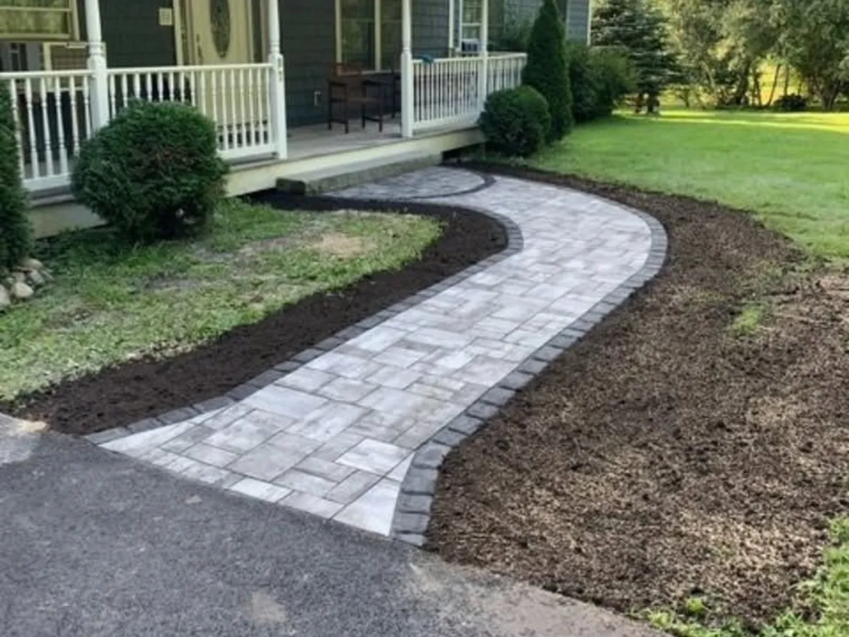 Curved gray paver walkway with a darker border leading to a covered front porch, with fresh soil beds on each side